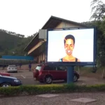 A front-facing view of an Adhaiwell P10 outdoor LED advertising billboard installed on a single pole in a parking lot in Rwanda, displaying a vibrant graphic during daylight with a warehouse and hills in the background.