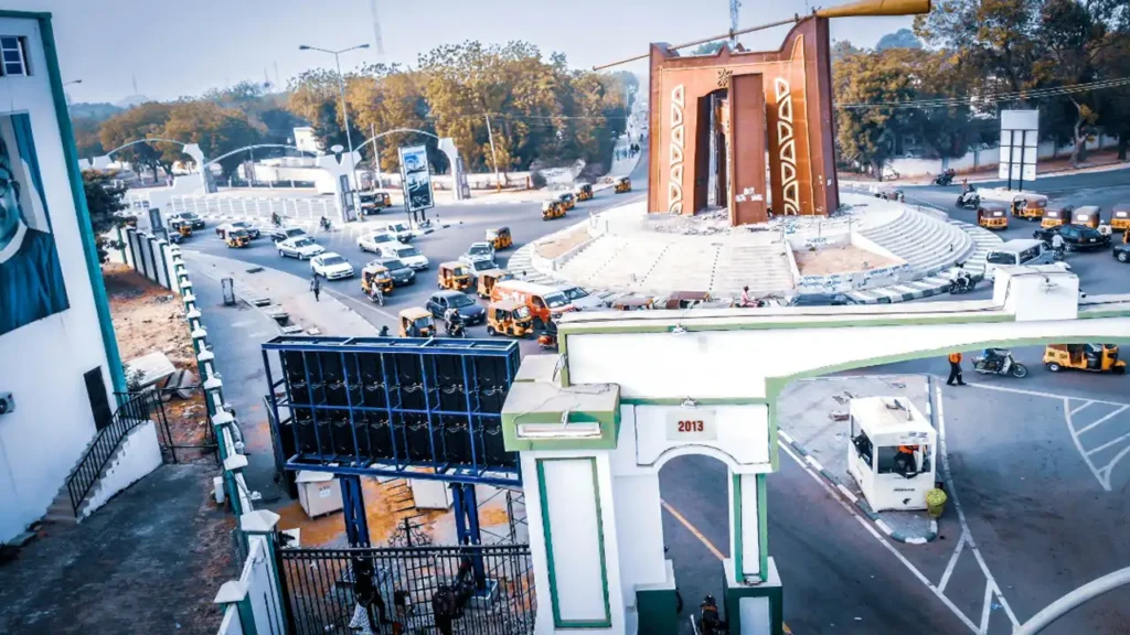 A massive Adhaiwell P10 die-cast aluminum LED display billboard installed on a multi-sided structure overlooking a busy traffic roundabout with yellow tuk-tuks and cars in Lagos, Nigeria. The landmark Gani Fawehinmi Park gate with the date "2013" is visible.