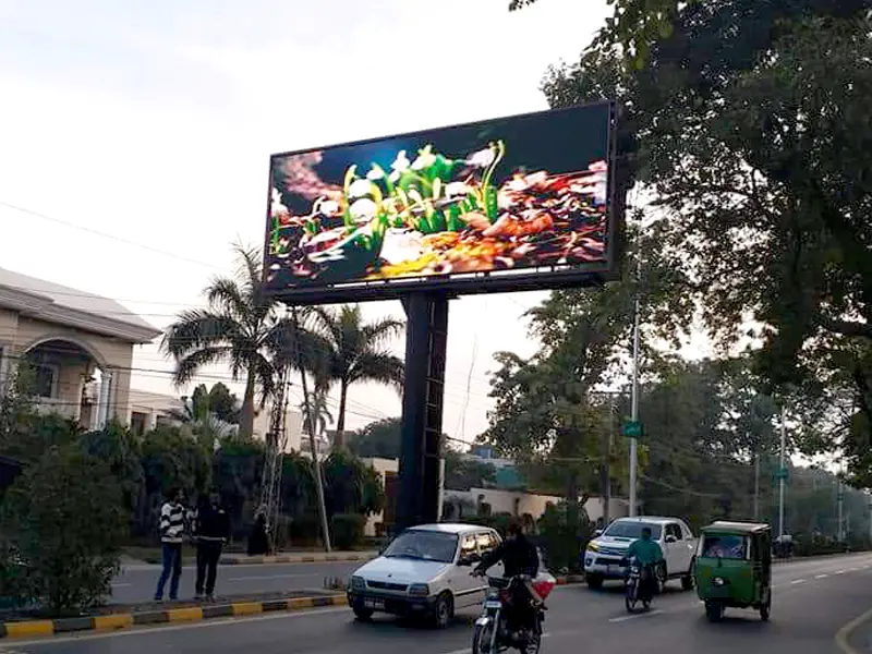 A vibrant, high-brightness Adhaiwell die-cast aluminum outdoor LED billboard installed on a single pole overlooking a busy street in Pakistan with a historic white mosque and rickshaws in the background.