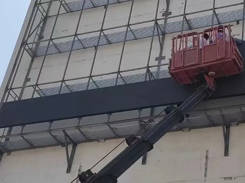 A construction worker in a hard hat on a Kasseler Hubwagen aerial platform platform lifts a die-cast aluminum LED display cabinet. It is being fixed onto a wall-mounted steel structure on a building in Dubai, UAE