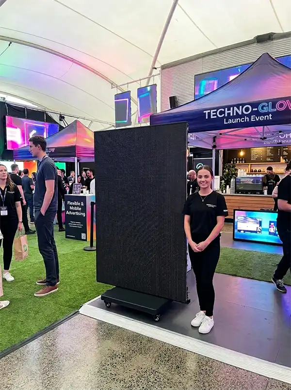 mobile LED poster screen on a rolling base stands next to a smiling female brand ambassador at a bustling indoor product launch event, with event tents, lighting, and other attendees in the background.